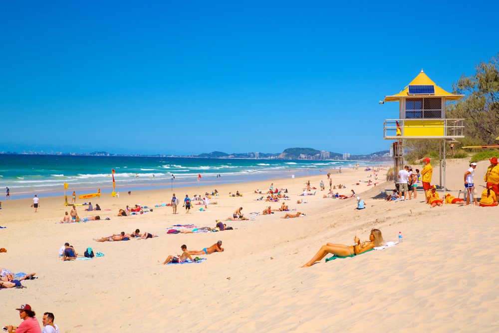 Beachgoers relax in the sunshine on a wide strip of Kurrawa Beach with a yellow lifeguard station in the background